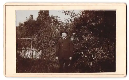 Fotografie unbekannter Fotograf und Ort, Portrait stattlicher Mann steht mit Hut im Anzug im Garten