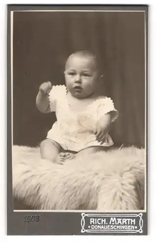 Fotografie Rich. Marth, Donaueschingen, Niedliches Kleinkind im weissen Kleidchen sitzt auf Teppich