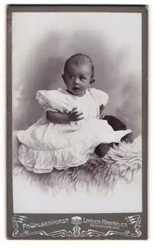 Fotografie Fr. Walkenhorst, Hannover-Linden, Portrait niedliches Kleinkind im weissen Kleid auf Fell sitzend