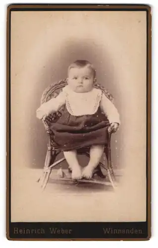 Fotografie Heinrich Weber, Winnenden, Portrait niedliches Kleinkind im hübschen Kleid mit Latz auf Korbstuhl sitzend