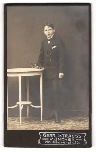Fotografie Gebr. Strauss, München, Portrait halbwüchsiger Knabe im Anzug mit Fliege und Buch