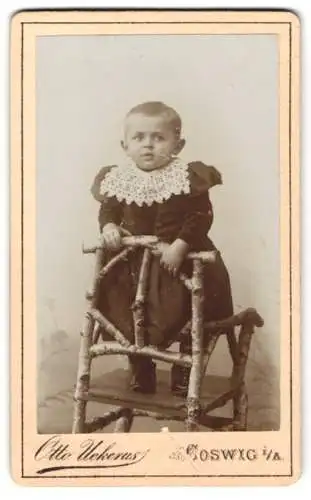 Fotografie Otto Uekerus, Coswig i. A., Süsses Baby im Kleid auf Birkenholz-Stuhl